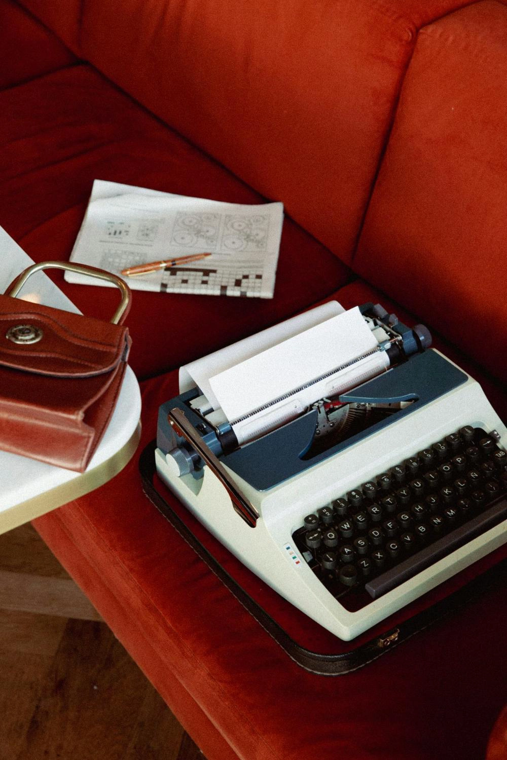 A vintage typewriter placed on a red couch. Next to the typewriter, there is a brown leather handbag on a white table. There is also a piece of paper with some printed text and drawings.