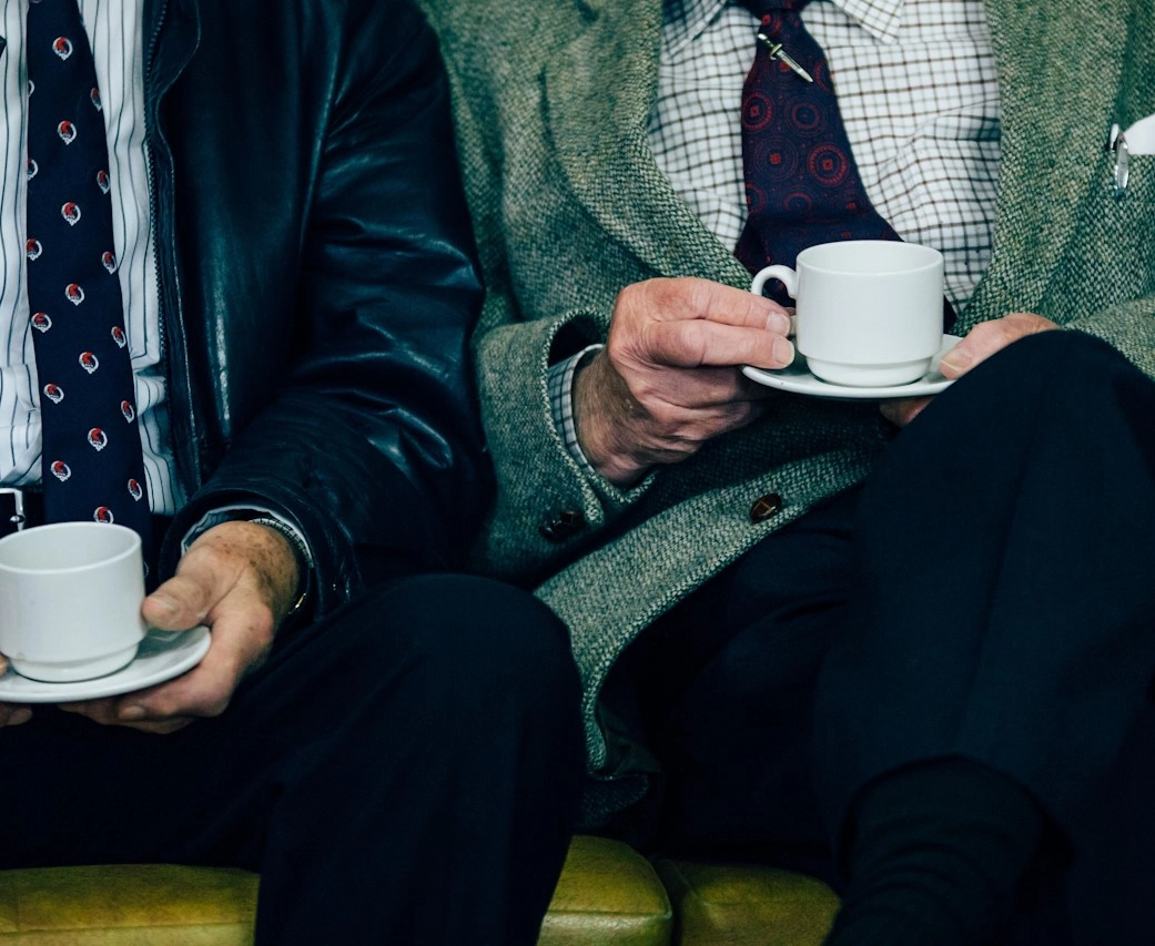Two elderly men sitting next to each other, holding white cups and saucers. Both are dressed in formal attire.