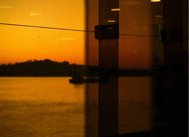 Sonnenuntergang über einem Gewässer. Im Vordergrund sind reflektierende Glasflächen zu sehen, die das Licht des Sonnenuntergangs einfangen. Im Hintergrund ist eine dunkle Silhouette einer Landschaft oder Insel zu erkennen.